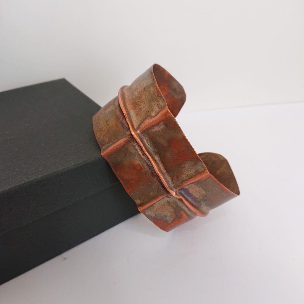Chunky copper cuff bracelet or bangle with aged copper colour patina and fold formed design, resting against its presentation box