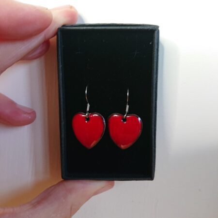 Red heart shaped enamelled earrings in their presentation box. The earrings have surgical steel earhooks. The red is a slightly darker than scarlet shade and a little dull rather than vivid.