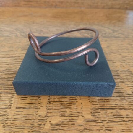 Copper wire bangle or bracelet with curled ends and slight overlap. The bracelet has an aged copper colour patina - between milk and dark chocolate brown. It also has a light hammered texture. This picture shows it resting on the lid of its black presentation box