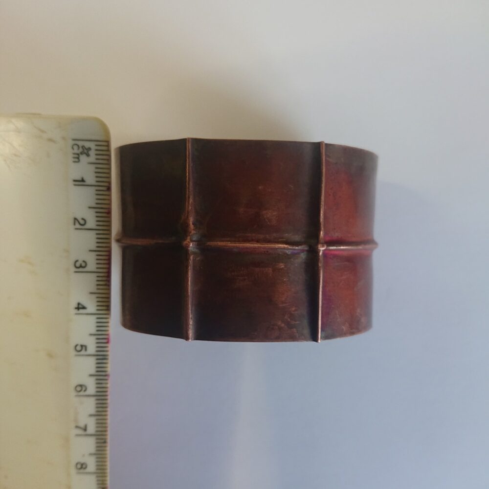 Copper bracelet or bangle with fold-formed ridges in a chunky cuff style. There is a centre ridge line, plus two vertical ridge or fold lines. It has an aged copper colour patina, so a mid to darker brown. This photo shows the bracelet next to a ruler to show its height is about 4cm
