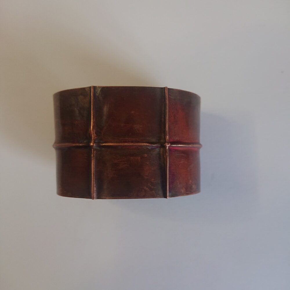 Copper bracelet or bangle with fold-formed ridges in a chunky cuff style. There is a centre ridge line, plus two vertical ridge or fold lines. It has an aged copper colour patina, so a mid to darker brown.