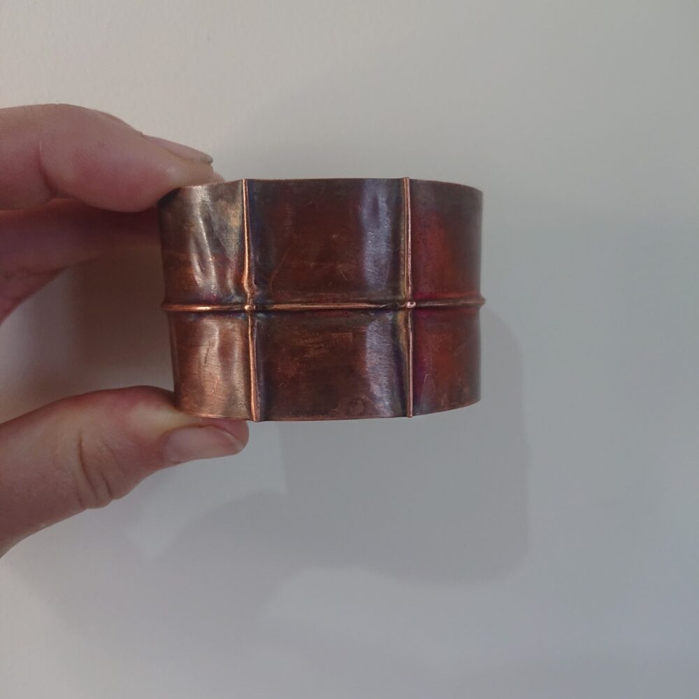 Copper bracelet or bangle with fold-formed ridges in a chunky cuff style. There is a centre ridge line, plus two vertical ridge or fold lines. It has an aged copper colour patina, so a mid to darker brown.