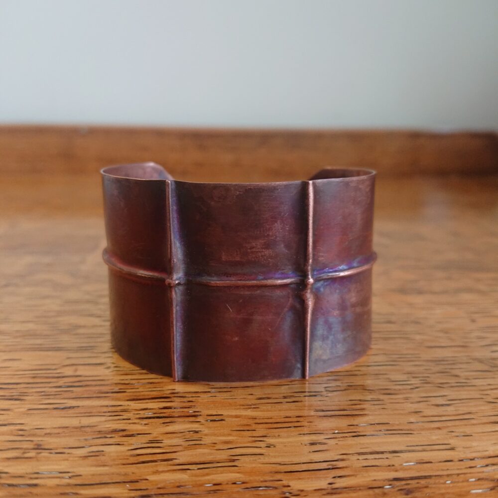 Copper bracelet or bangle with fold-formed ridges in a chunky cuff style. There is a centre ridge line, plus two vertical ridge or fold lines. It has an aged copper colour patina, so a mid to darker brown.