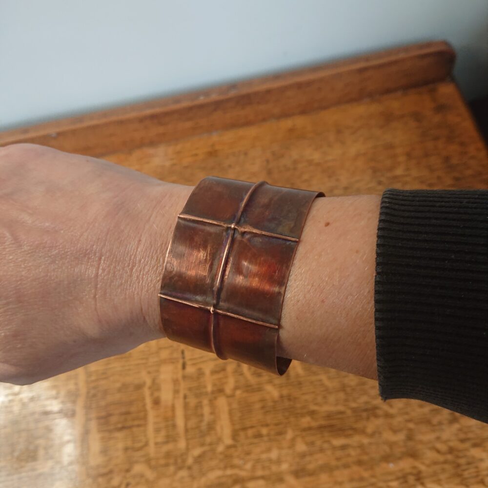 Copper bracelet or bangle with fold-formed ridges in a chunky cuff style. There is a centre ridge line, plus two vertical ridge or fold lines. It has an aged copper colour patina, so a mid to darker brown. This photo shows me wearing the bracelet