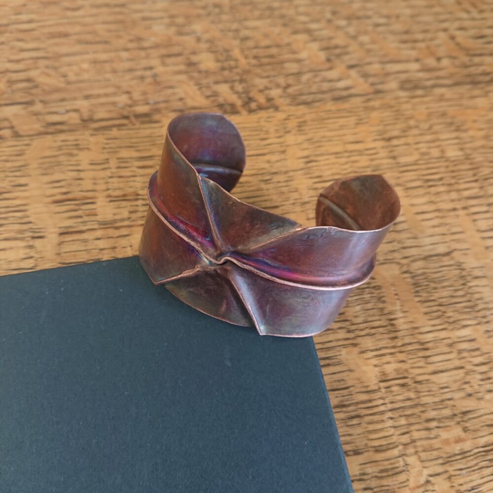 Copper bracelet or bangle with fold-formed ridges in a chunky cuff style. There is a centre ridge line, plus two crossing diagonal ridges or fold lines. It has an aged copper colour patina, so a mid to darker brown. This photo shows the bracelet resting on the lid of its black presentation box