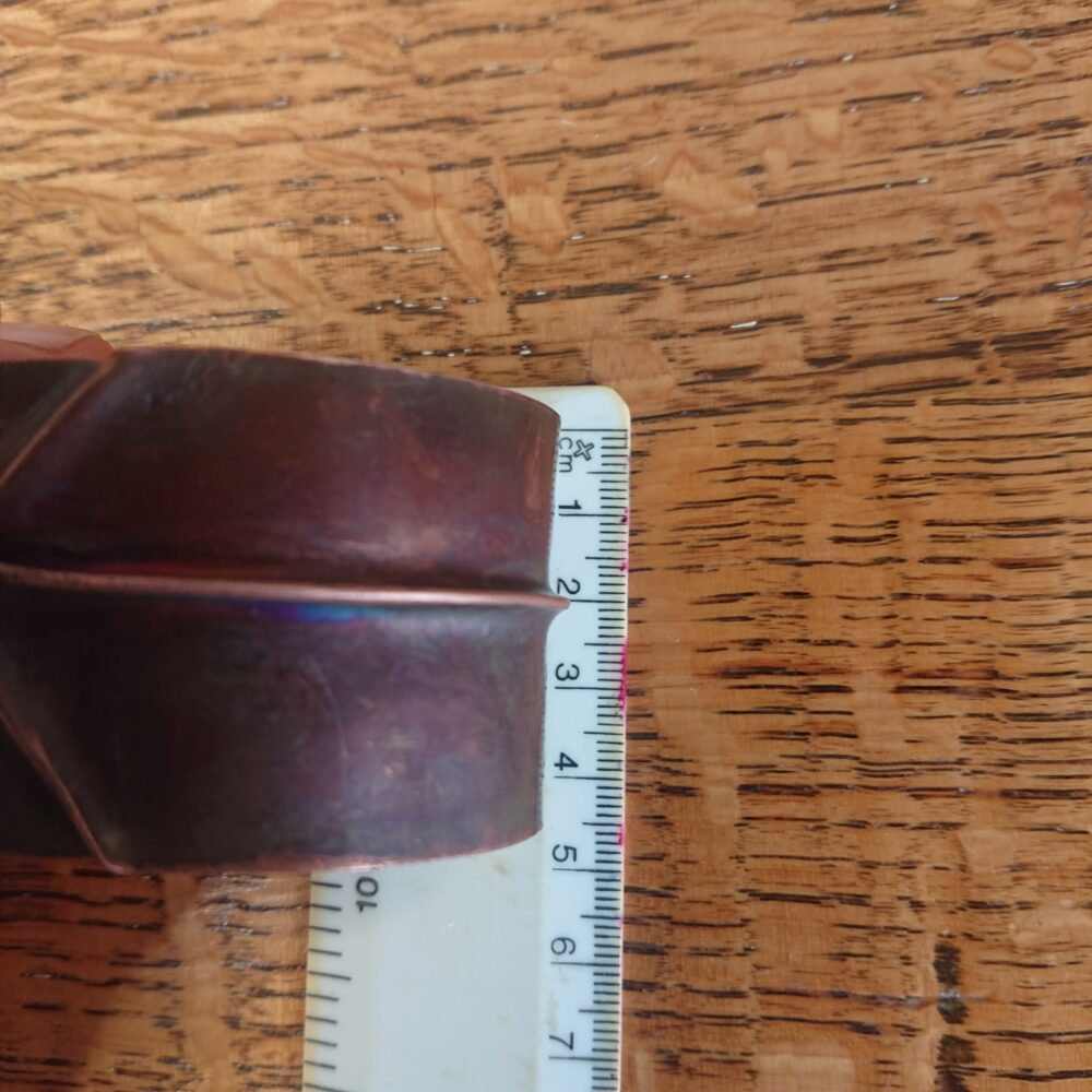 Copper bracelet or bangle with fold-formed ridges in a chunky cuff style. There is a centre ridge line, plus two crossing diagonal ridges or fold lines. It has an aged copper colour patina, so a mid to darker brown. This photo shows the bracelet beside a ruler to show it's about 4cm tall
