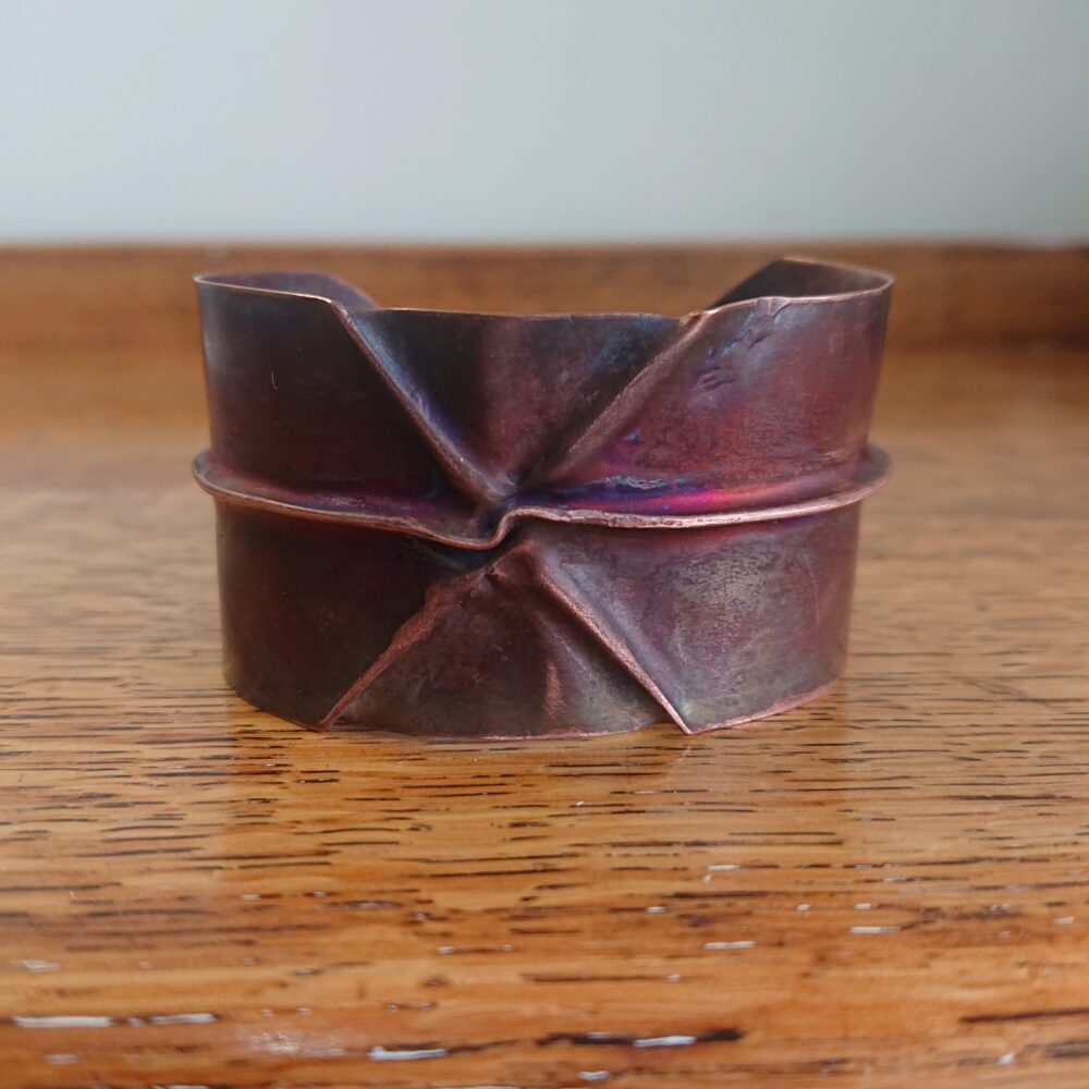 Copper bracelet or bangle with fold-formed ridges in a chunky cuff style. There is a centre ridge line, plus two crossing diagonal ridges or fold lines. It has an aged copper colour patina, so a mid to darker brown.