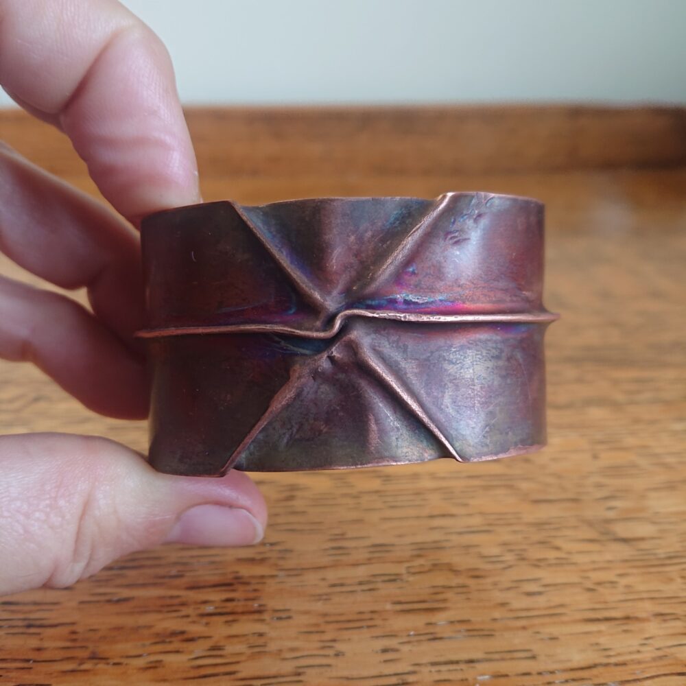 Copper bracelet or bangle with fold-formed ridges in a chunky cuff style. There is a centre ridge line, plus two crossing diagonal ridges or fold lines. It has an aged copper colour patina, so a mid to darker brown.