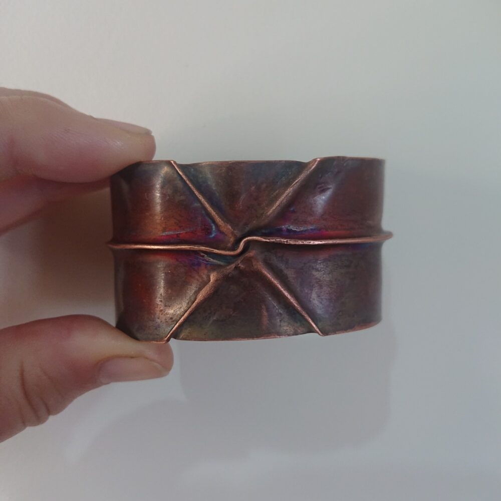 Copper bracelet or bangle with fold-formed ridges in a chunky cuff style. There is a centre ridge line, plus two crossing diagonal ridges or fold lines. It has an aged copper colour patina, so a mid to darker brown.