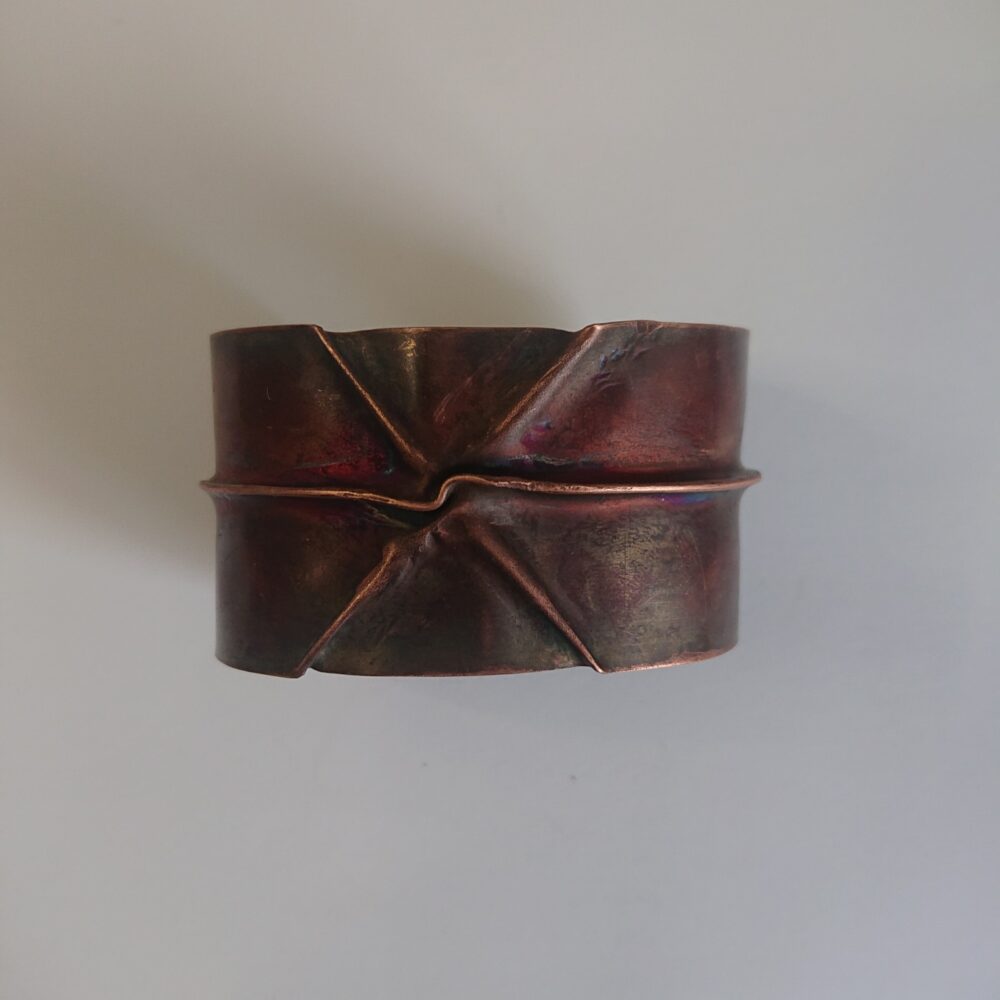 Copper bracelet or bangle with fold-formed ridges in a chunky cuff style. There is a centre ridge line, plus two crossing diagonal ridges or fold lines. It has an aged copper colour patina, so a mid to darker brown.