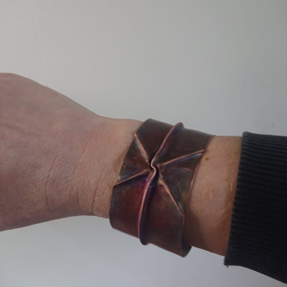 Copper bracelet or bangle with fold-formed ridges in a chunky cuff style. There is a centre ridge line, plus two crossing diagonal ridges or fold lines. It has an aged copper colour patina, so a mid to darker brown. This photo shows me wearing the bracelet