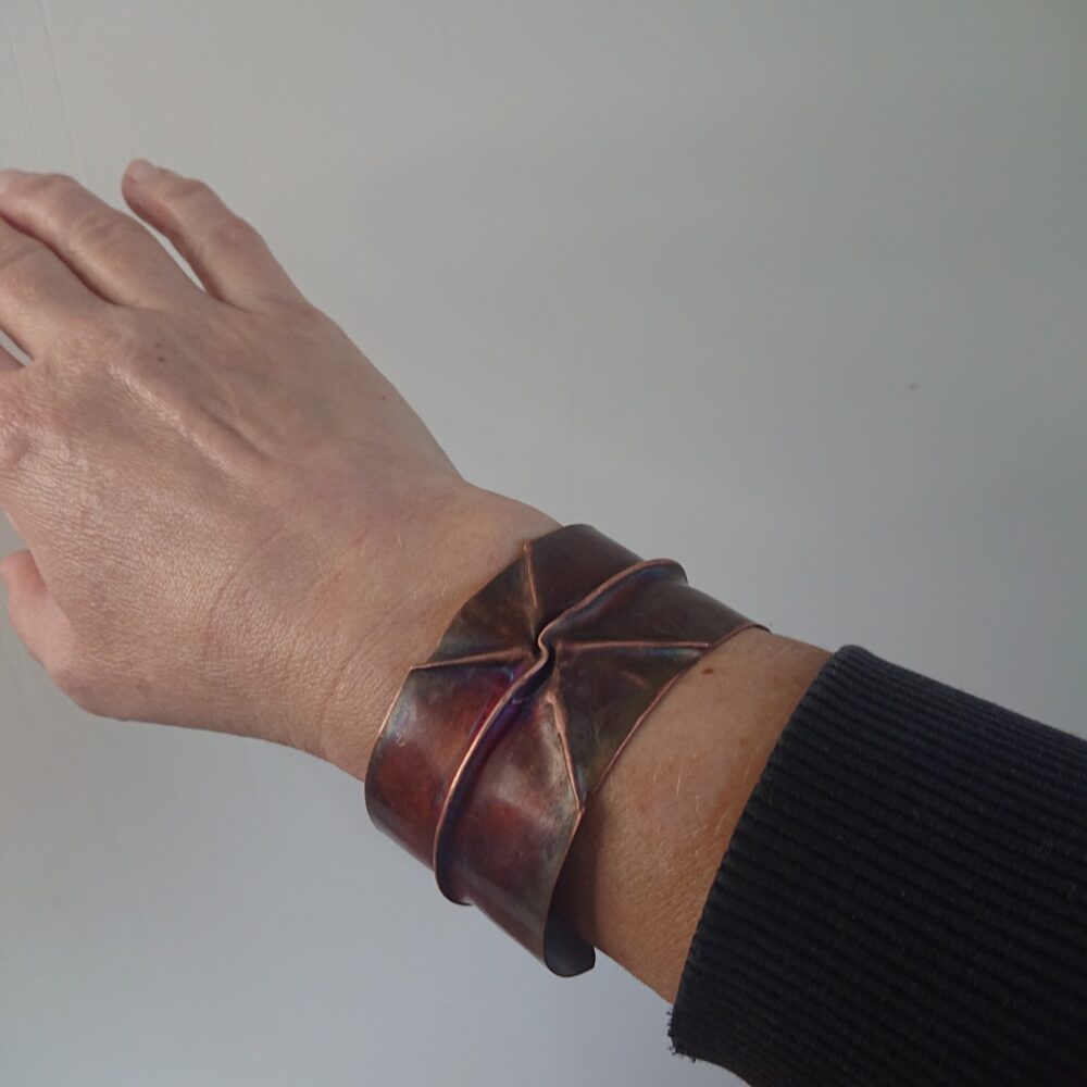 Copper bracelet or bangle with fold-formed ridges in a chunky cuff style. There is a centre ridge line, plus two crossing diagonal ridges or fold lines. It has an aged copper colour patina, so a mid to darker brown. This photo shows me wearing the bracelet