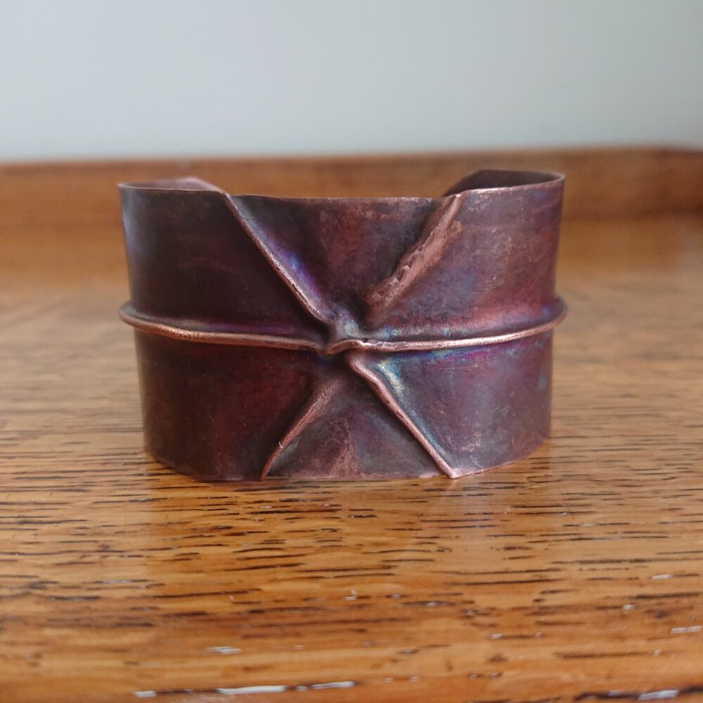 Copper bracelet or bangle with fold-formed ridges in a chunky cuff style. There is a centre ridge line, plus two crossing diagonal ridges or fold lines. It has an aged copper colour patina, so a mid to darker brown.