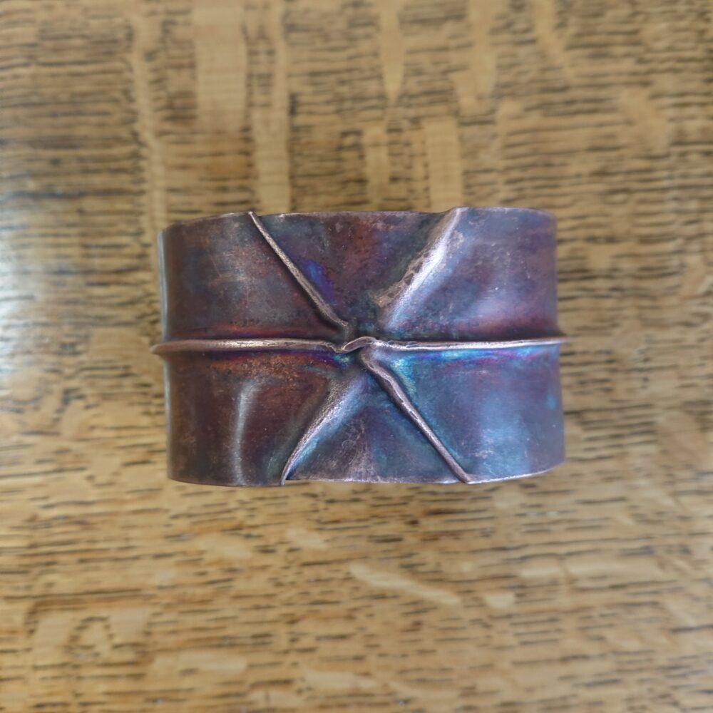Copper bracelet or bangle with fold-formed ridges in a chunky cuff style. There is a centre ridge line, plus two crossing diagonal ridges or fold lines. It has an aged copper colour patina, so a mid to darker brown.
