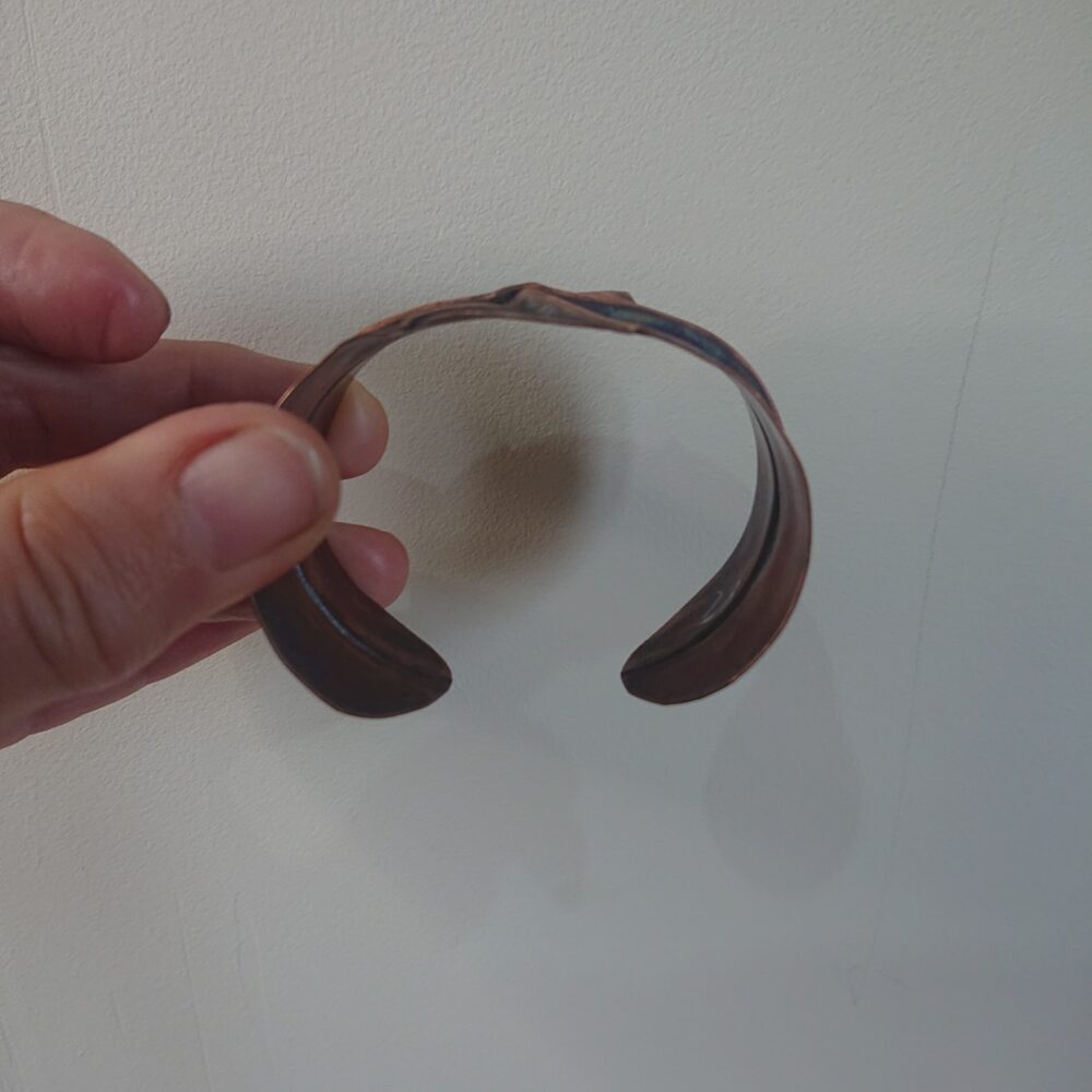 Copper bracelet or bangle with fold-formed ridges in a chunky cuff style. There is a centre ridge line, plus two crossing diagonal ridges or fold lines. It has an aged copper colour patina, so a mid to darker brown. The style is perhaps bohemian or industrial. It is unisex.