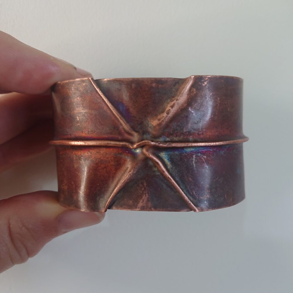 Copper bracelet or bangle with fold-formed ridges in a chunky cuff style. There is a centre ridge line, plus two crossing diagonal ridges or fold lines. It has an aged copper colour patina, so a mid to darker brown. The style is perhaps bohemian or industrial. It is unisex.