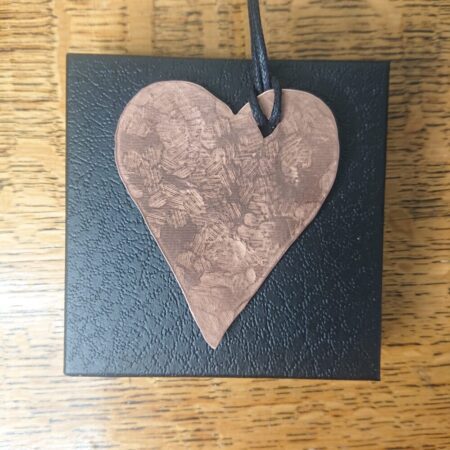 Heart shaped copper necklace with hammer texture marks. The necklace has a black waxed cotton cord, through a heart shaped hole and comes in a black presentation box. The copper is bright, shiny copper, protected with lacquer. The pendant is about 7cm tall and about 5.5cm wide at the widest point. It has a slight dome or curve to it.