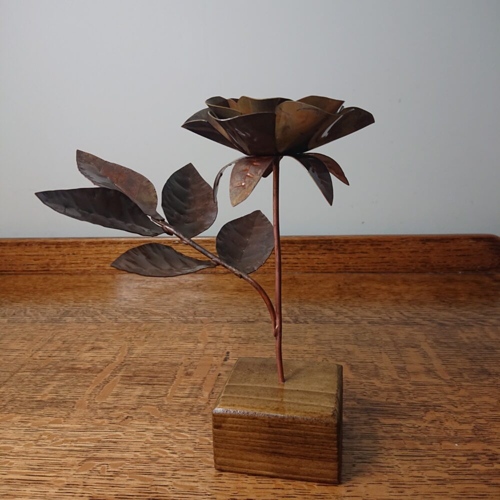 Handmade copper rose in a dark, aged copper shade with a wooden square base. The rose has an open flower on an upright stem and leaves on a side stem. It's about 19cm tall