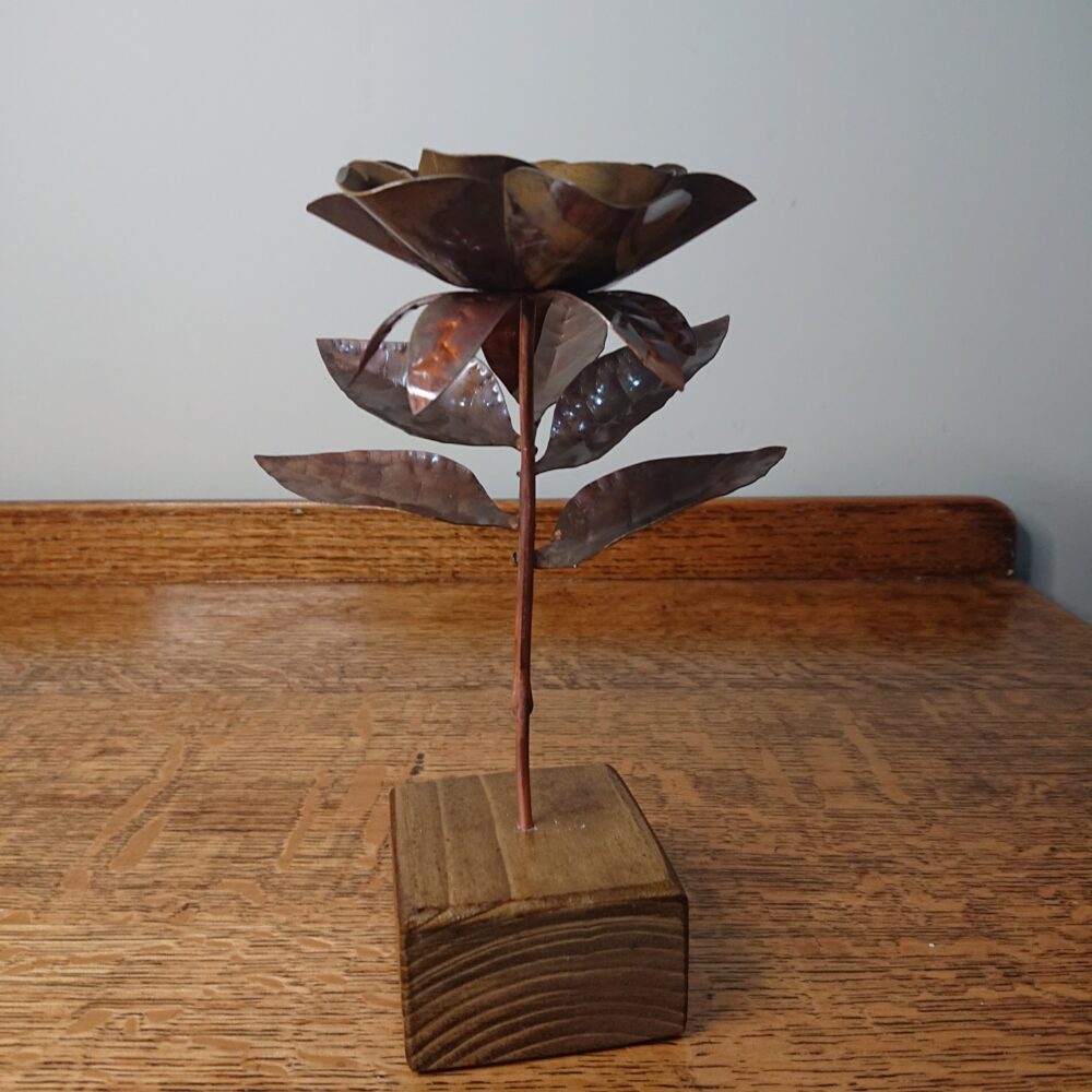 Handmade copper rose in a dark, aged copper shade with a wooden square base. The rose has an open flower on an upright stem and leaves on a side stem. It's about 19cm tall