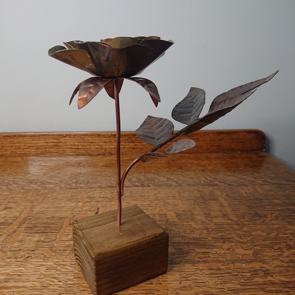 Handmade copper rose in a dark, aged copper shade with a wooden square base. The rose has an open flower on an upright stem and leaves on a side stem.