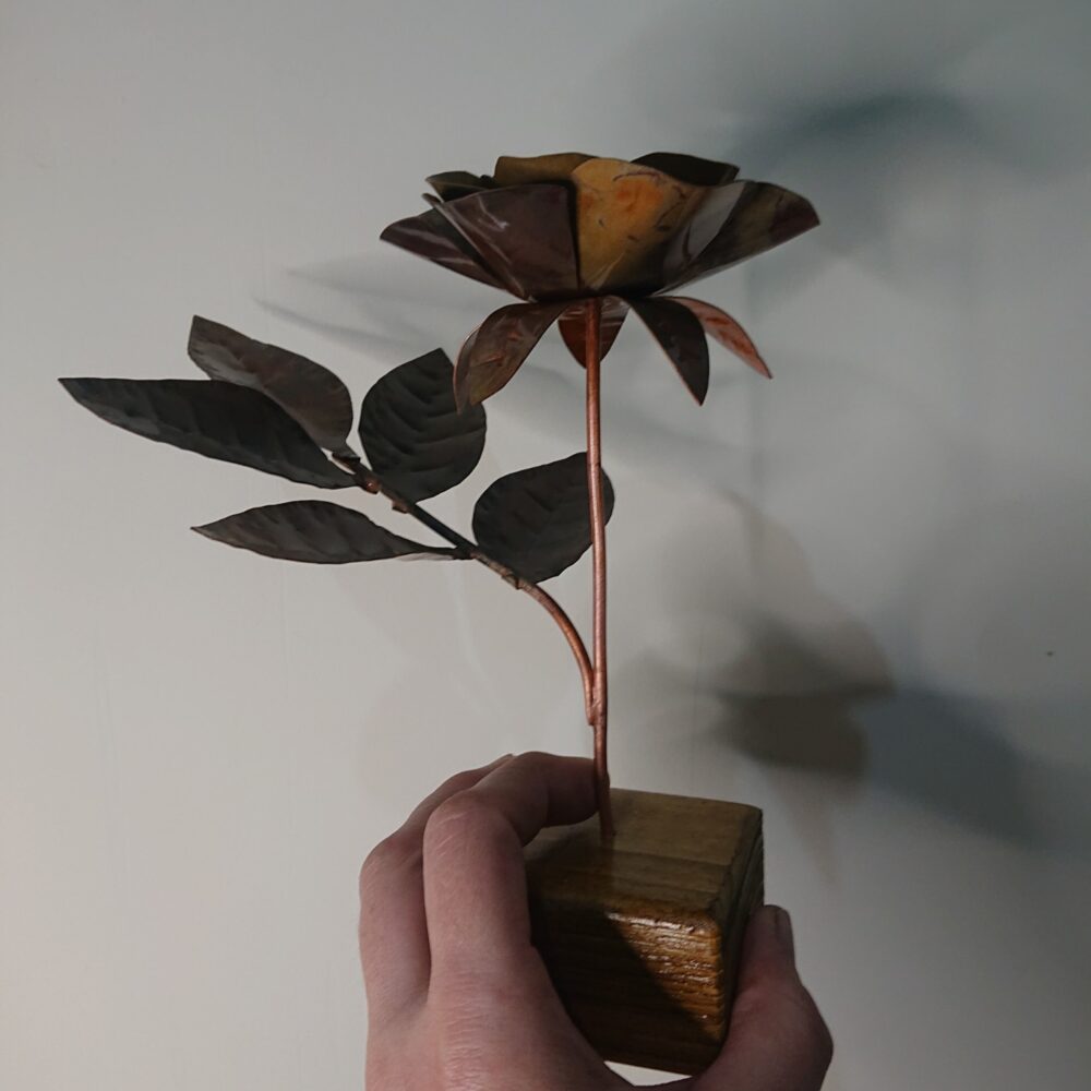 Handmade copper rose in a dark, aged copper shade with a wooden square base. The rose has an open flower on an upright stem and leaves on a side stem.