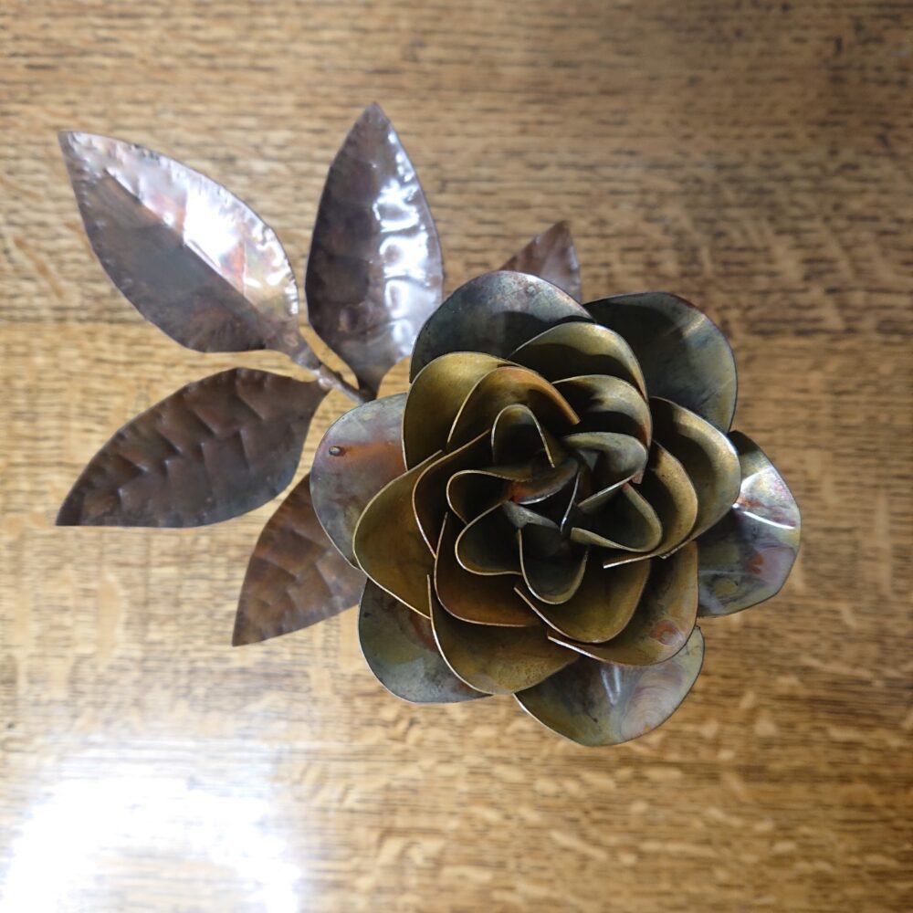 Handmade copper rose in a dark, aged copper shade with a wooden square base. The rose has an open flower on an upright stem and leaves on a side stem. This is the view from above