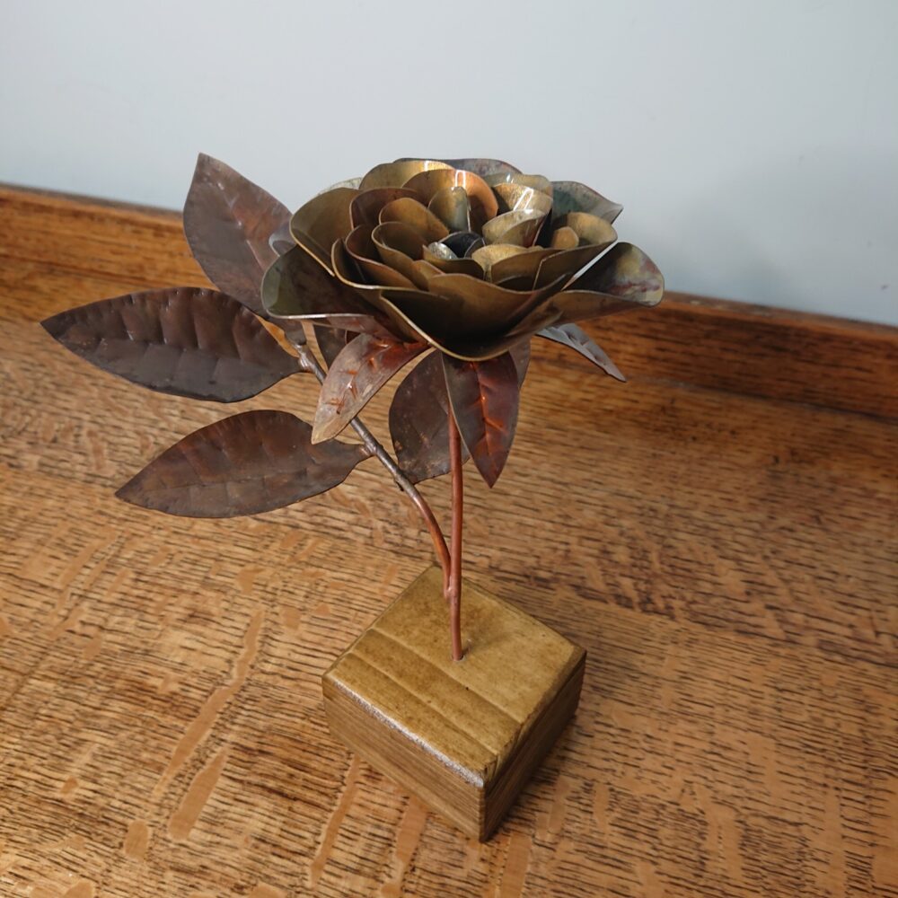 Handmade copper rose in a dark, aged copper shade with a wooden square base. The rose has an open flower on an upright stem and leaves on a side stem.