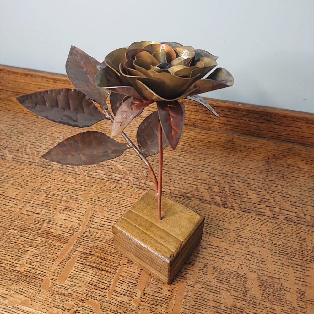 Handmade copper rose in a dark, aged copper shade with a wooden square base. The rose has an open flower on an upright stem and leaves on a side stem.