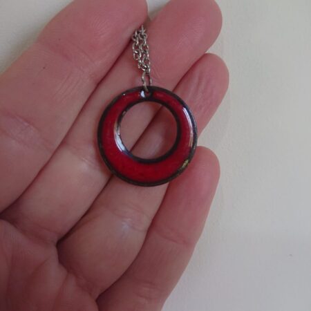 Red round enamelled necklace pendant with silver coloured chain. The pendant is a circle or round, with a round hole in it, slightly offset towards the top. I overfired the red so it has dark dots in it and a darker outer edge. The edge and dots look black.