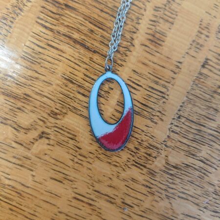 Oval shaped necklace pendant with an oval hole in it. The pendant is pale grey, with a red section near the bottom. The pendant has a slightly darker outer edge and tiny darker dots in it. It has a silver coloured chain.