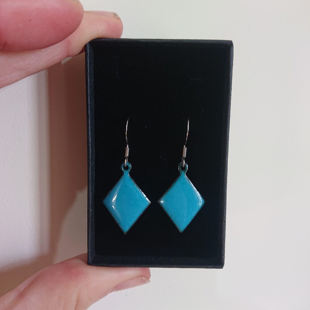 Teal diamond or trapezoid shaped earrings with ear hooks and in a black presentation box. The teal colour is turquoise blue to my eyes. I fired them for longer to give tiny darker dots to make them more interesting.