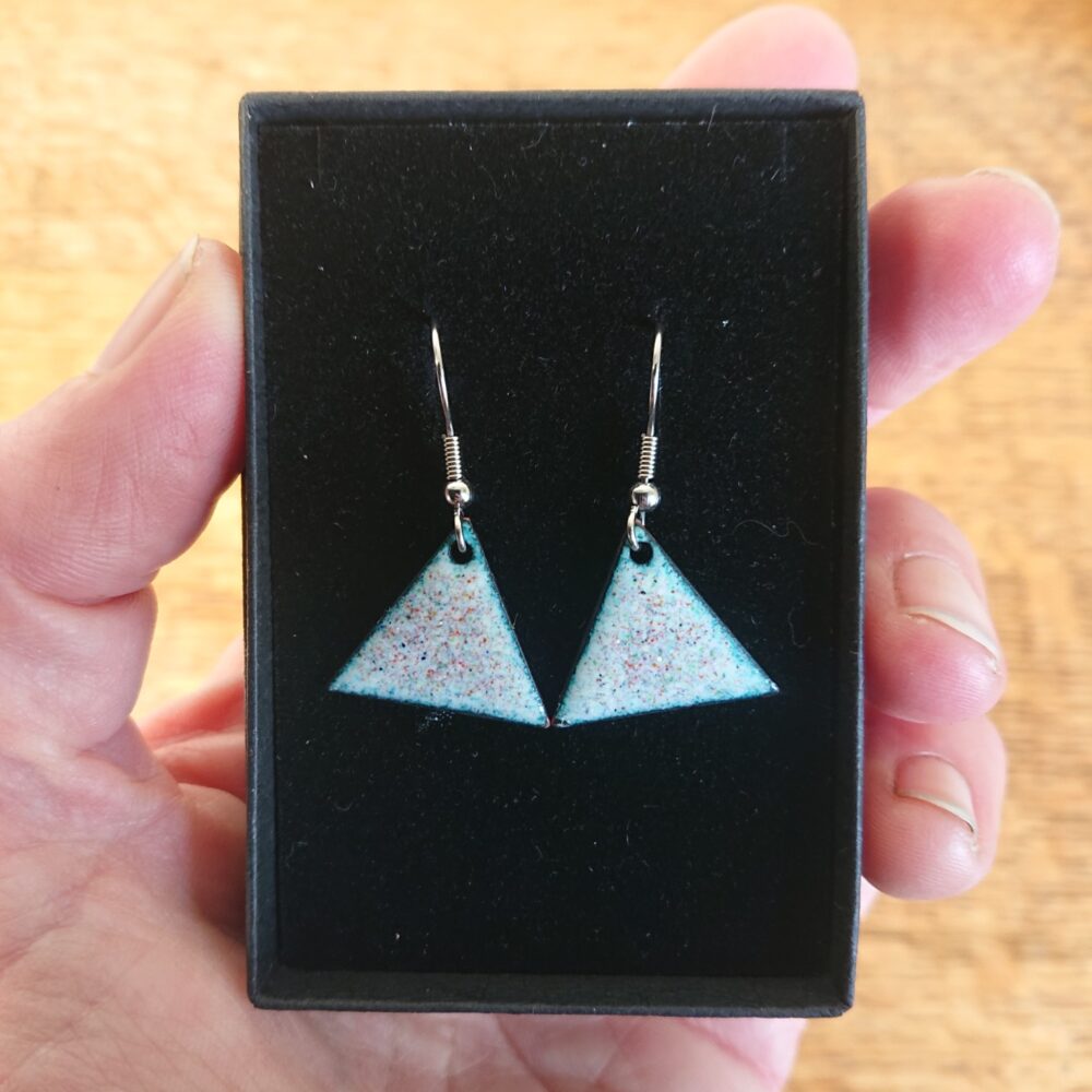 Triangle or triangular shaped earrings with ear hooks. The earrings have pale backgrounds with lots of tiny coloured dots or spots. They are shown in their black presentation box.