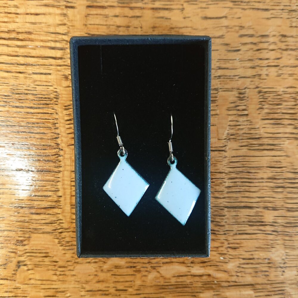 Light grey with a lilac tint enamelled copper earrings. The earrings have been fired for longer so have dark dots. They're on ear hooks. The shape is diamond or trapezoid. On this picture they are in their presentation box.