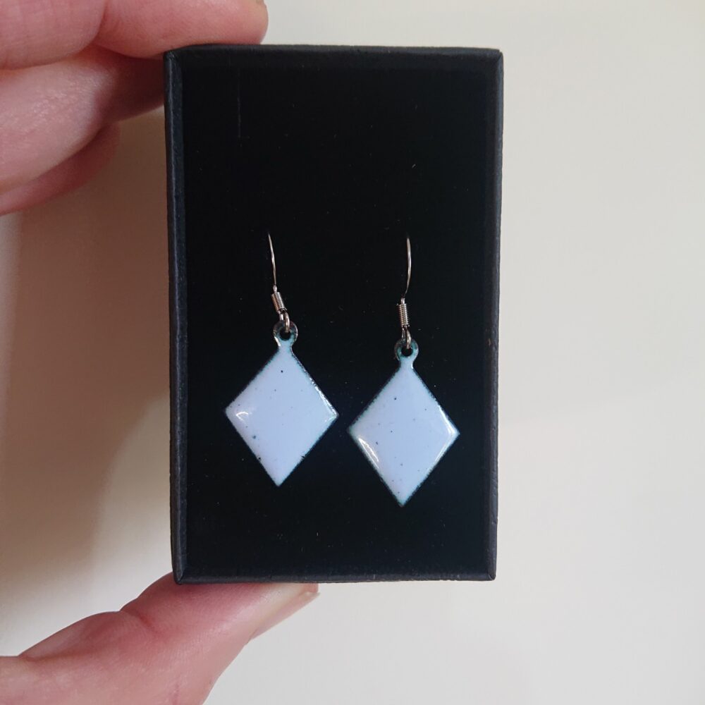 Light grey with a lilac tint enamelled copper earrings. The earrings have been fired for longer so have dark dots. They're on ear hooks. The shape is diamond or trapezoid. On this picture they are in their presentation box.