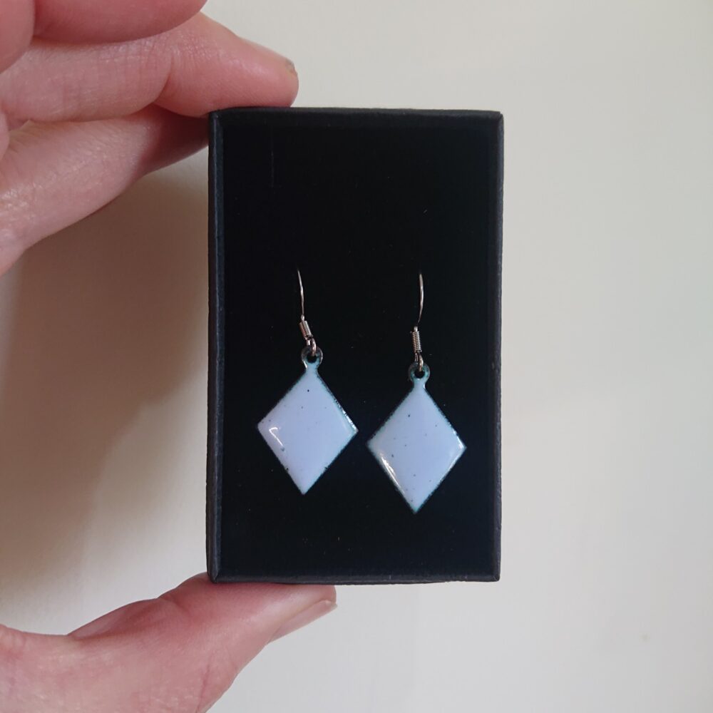 Light grey with a lilac tint enamelled copper earrings. The earrings have been fired for longer so have dark dots. They're on ear hooks. The shape is diamond or trapezoid. On this picture they are in their presentation box.