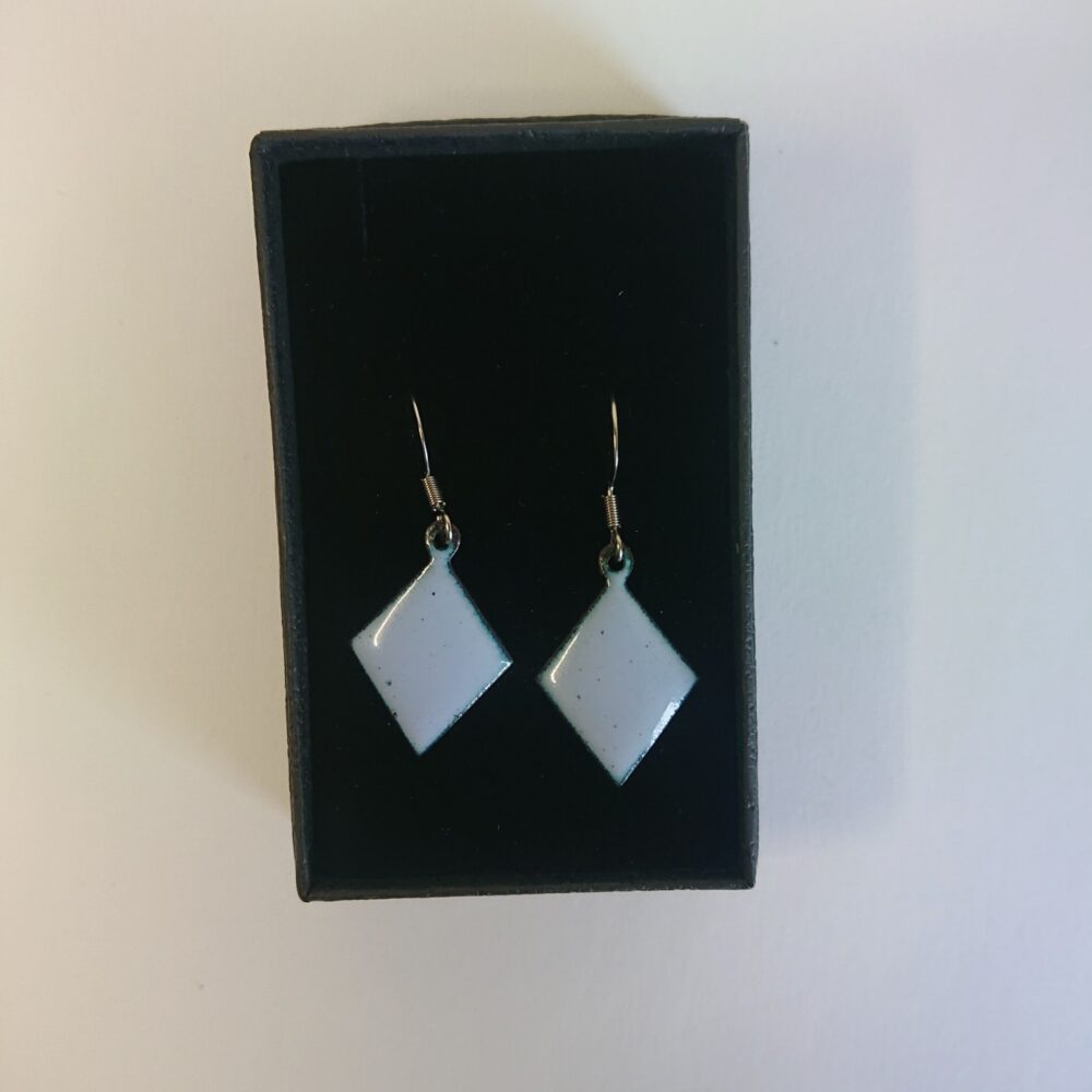 Light grey with a lilac tint enamelled copper earrings. The earrings have been fired for longer so have dark dots. They're on ear hooks. The shape is diamond or trapezoid. On this picture they are in their presentation box.