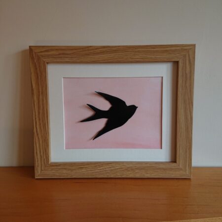 Enamelled bird wall art or wall hanging. The bird is black or a silhouette. The bird is meant to be a swallow, but it also looks a little like a swift or house martin. It is in flight. Behind the swallow is a peachy colour water colour background, which is slightly darker towards the bottom. The swallow is mounted into an oak effect frame. It is landscape orientation