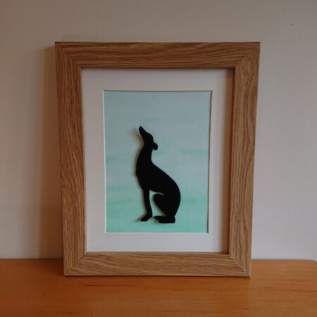 Enamelled greyhound wall art or wall hanging. The greyhound is black or a silhouette and is sitting, looking upwards. Behind the greyhound or whippet is a green gradient water colour background, with the darkest green at the bottom. The greyhound is mounted into an oak effect frame.